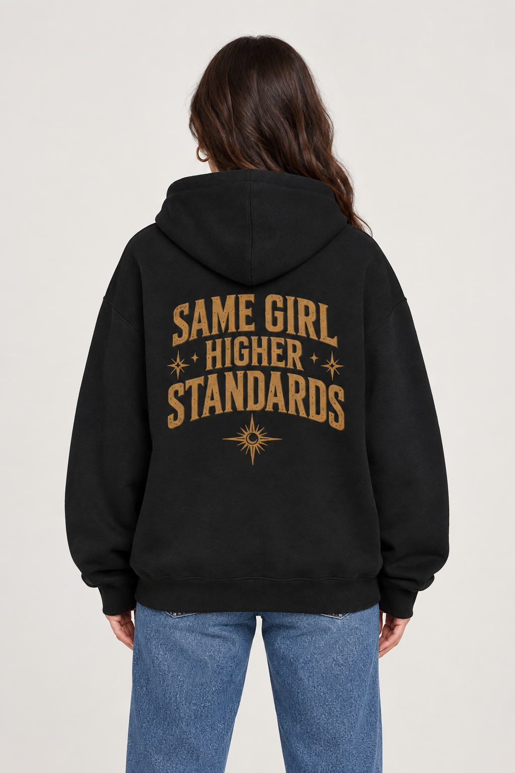 Woman wearing a black oversized hoodie with gold Same Girl Higher Standards lettering, stars and a sunburst on the back.